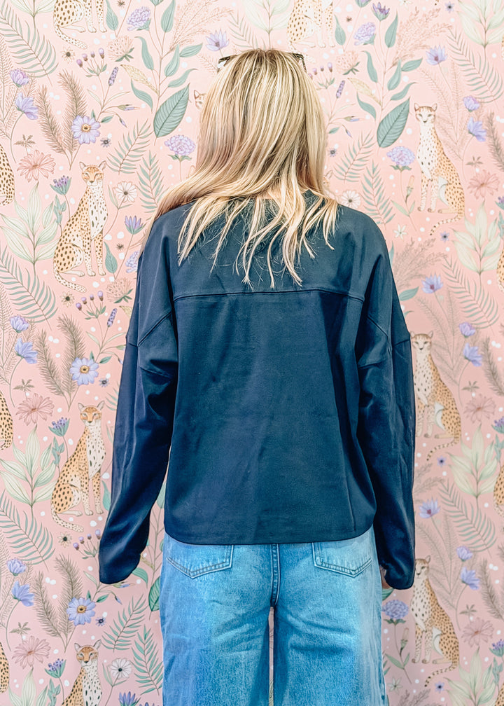 A woman viewed from behind wearing a dark navy blue long-sleeve shirt and light blue jeans, standing in front of a pink floral wallpaper with animal prints.