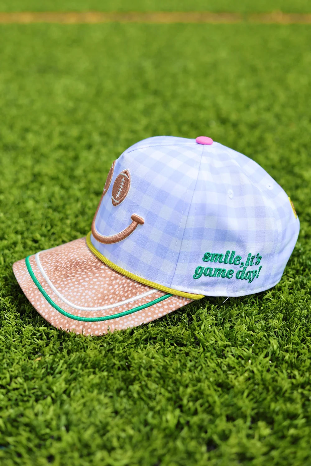 Blue gingham trucker hate with 'smile, it's game day!' embroidery on side sitting on grass