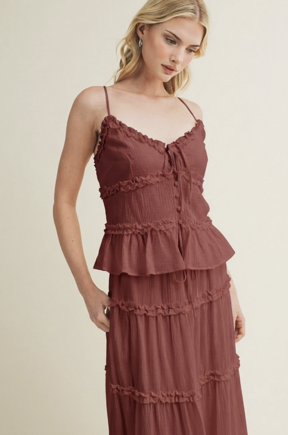 A woman wearing a rust-brown tiered spaghetti-strap dress with a ruffled bodice and tie-front detail, photographed against a plain light beige studio background.