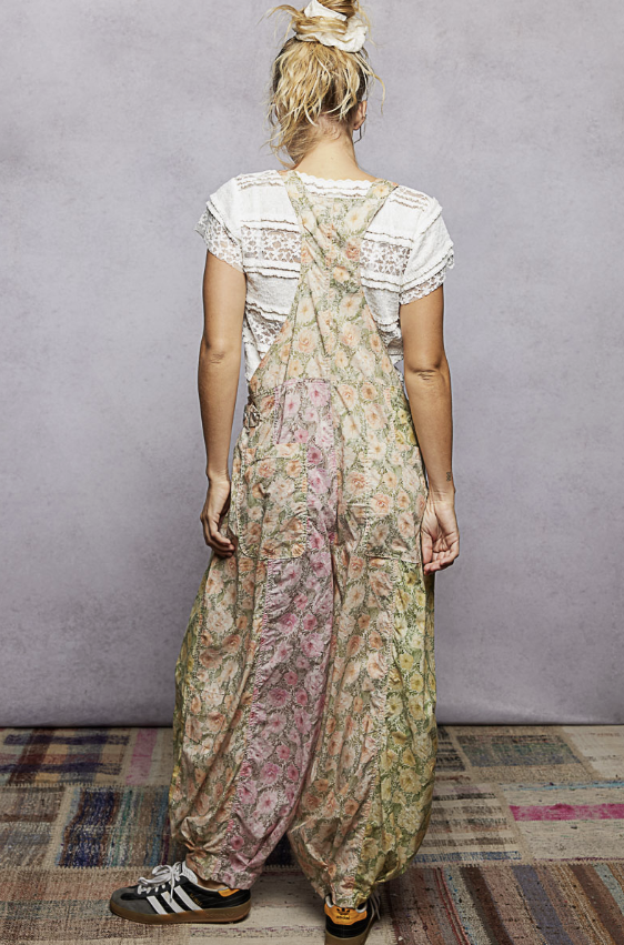 A woman shown from behind wearing a pair of loose, wide-leg floral print overall jumpsuits over a white lace-trim t-shirt, standing against a textured gray wall.