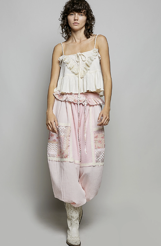 A model wearing a bohemian-style two-piece outfit consisting of a cream ruffled camisole top and light pink patchwork wide-leg pants with lace trim, paired with white lace-up boots, against a neutral gray background.