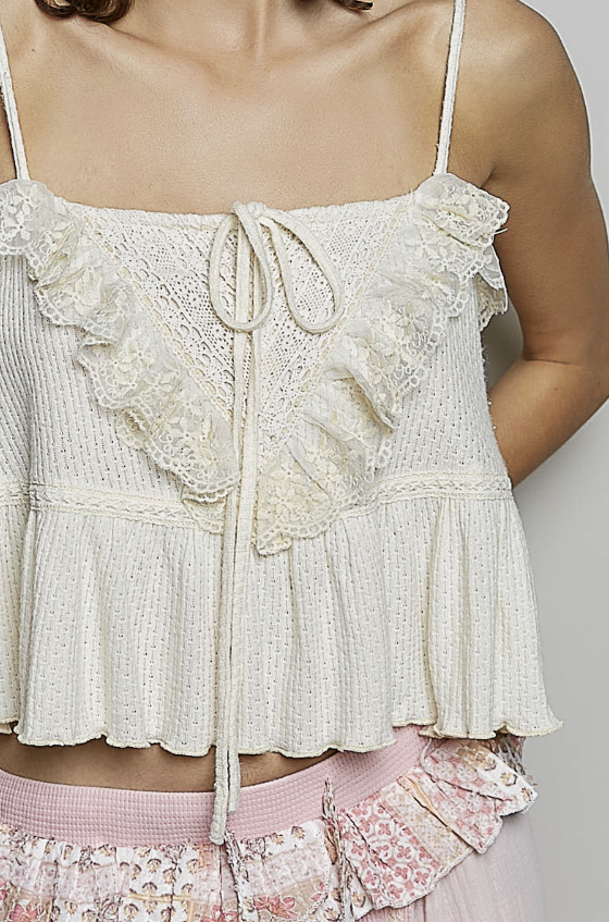 An off-white knit camisole top with ruffled lace trim, spaghetti straps, and a front tie closure, shown on a model.