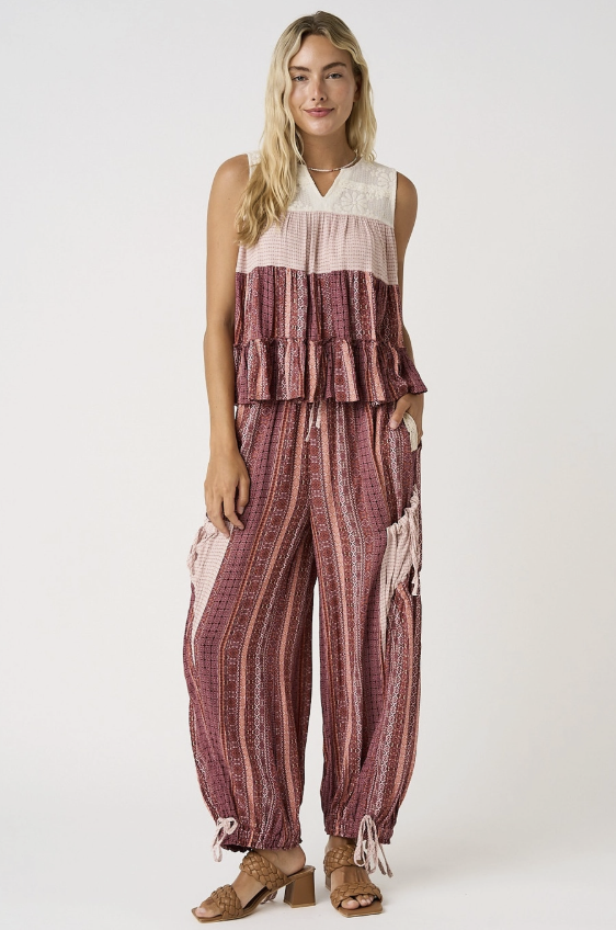 A woman modeling a sleeveless two-piece outfit with a cream ruffled top and matching wide-leg pants in a dusty rose and burgundy striped pattern, paired with brown heeled sandals against a plain white studio background.