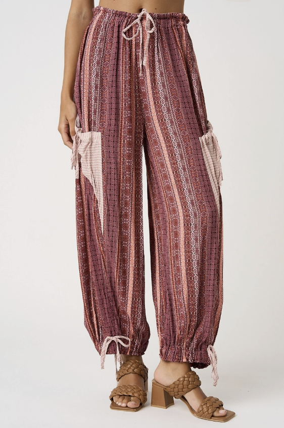 A person wearing loose, high-waisted patterned pants in dusty rose, burgundy, and cream, with drawstring waist and side tie details, paired with brown strappy heeled sandals against a light gray studio background.