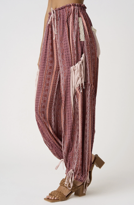 A person wearing loose, high-waisted harem-style pants in a dusty mauve and pink plaid pattern with cream fringe details, paired with tan heeled sandals against a plain light background.
