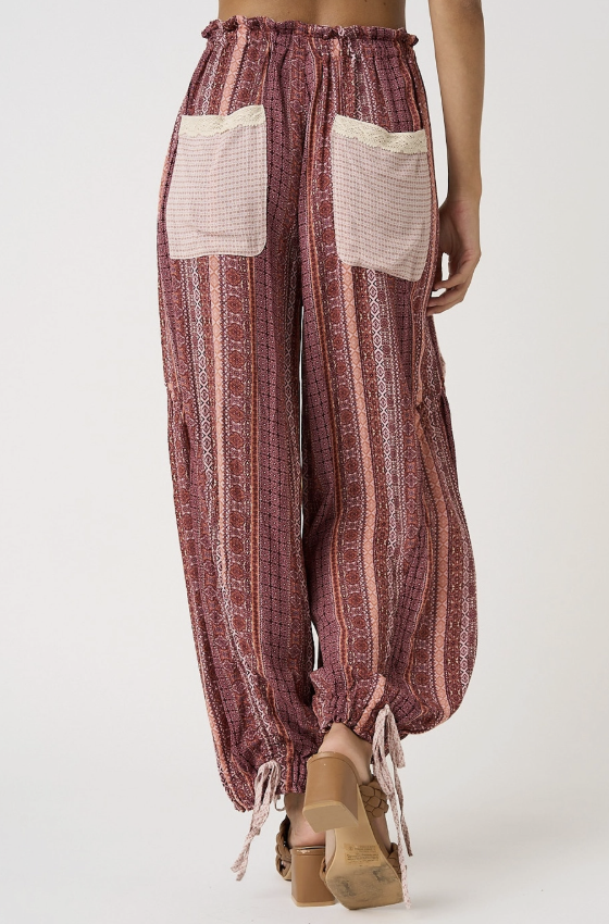 A pair of loose, high-waisted bohemian-style pants in a dusty rose and beige striped pattern, featuring two large cream mesh back patch pockets and drawstring hems, shown on a model from the back.