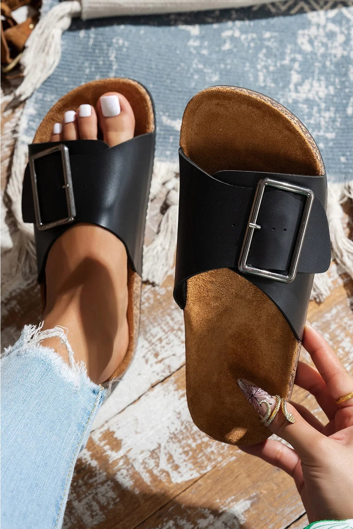 Black and brown sandals with buckles worn by a person on a textured surface.