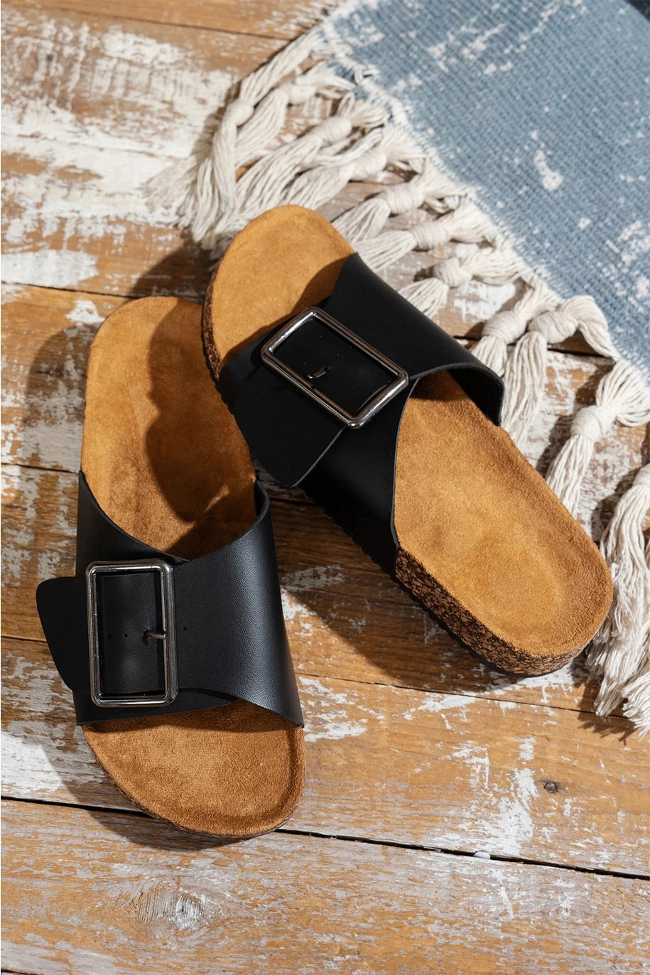 Pair of cork sandals with black buckles on a wooden surface.