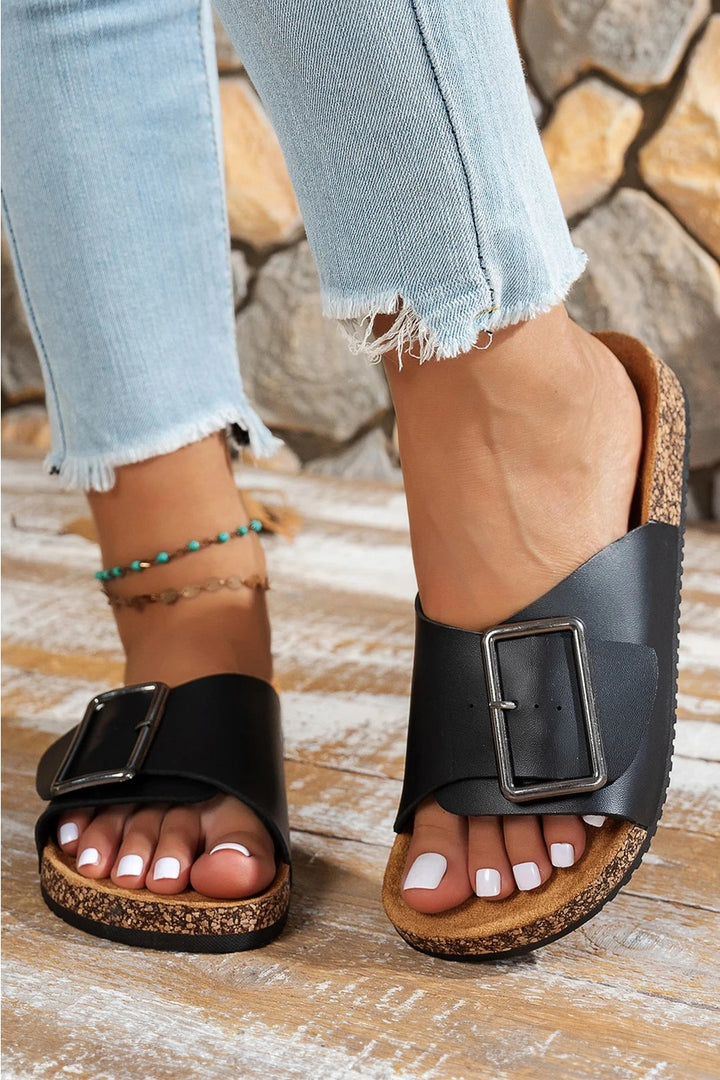 Black buckled sandals worn with light blue jeans on a wooden floor.