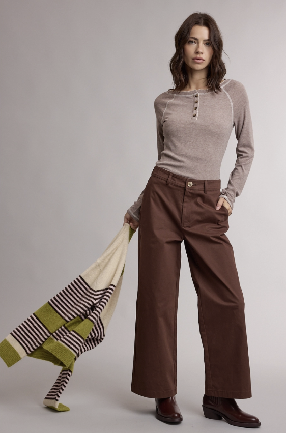 A woman stands against a light gray studio background wearing a fitted taupe henley top, brown wide-leg pants, and dark brown leather boots. She holds a cream knit sweater with green and black stripes in one hand.