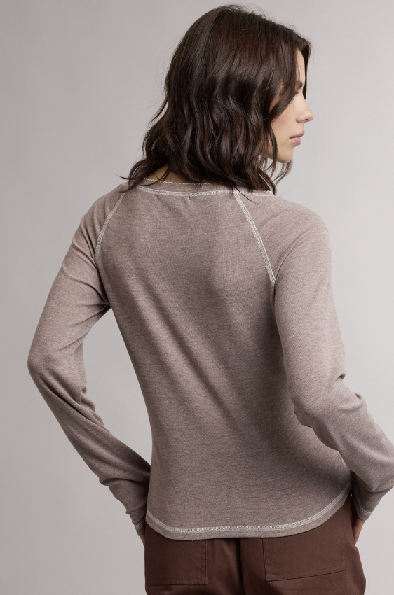 A woman shown from the back wearing a taupe heathered long-sleeve top with raglan sleeves and brown pants, photographed against a neutral light gray studio background.