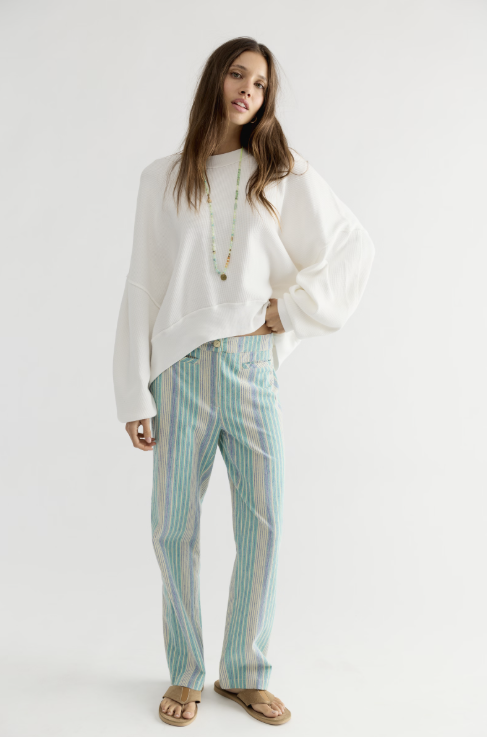 A woman modeling a white oversized sweatshirt and light blue and white vertically striped trousers, standing against a plain white studio background.