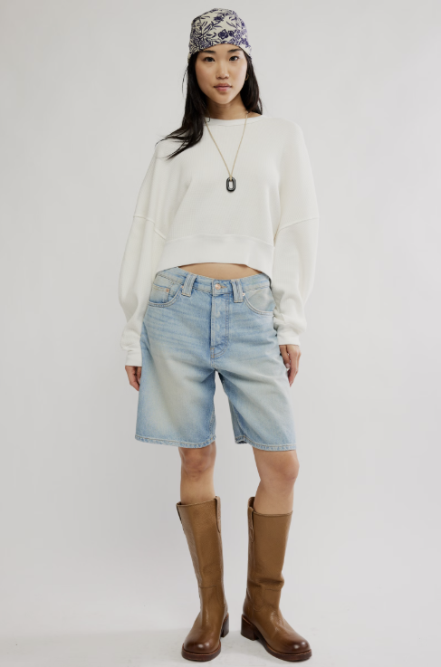 A model stands against a light gray studio background wearing a white cropped sweatshirt, light-wash denim shorts, brown knee-high boots, a patterned headband, and a necklace.