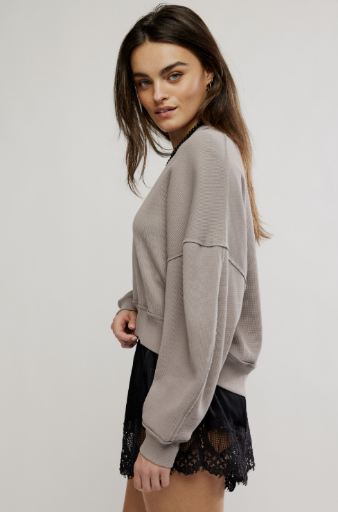 A woman wearing a taupe gray waffle-knit cropped sweatshirt and black lace shorts, photographed against a light gray studio background.