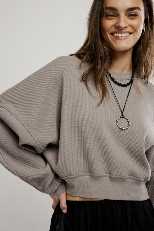 A woman wearing a taupe ribbed knit crewneck sweatshirt with oversized balloon sleeves, styled with black accessories against a light gray background.