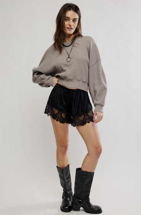 A model wearing a taupe cropped sweatshirt, black pleated mesh shorts with a lace hem, and black knee-high boots, standing against a light gray studio background.