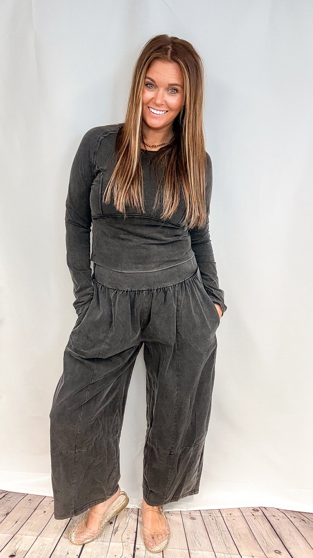 A woman modeling a dark charcoal gray two-piece lounge set consisting of a long-sleeve top and matching wide-leg pants, standing against a white studio backdrop.