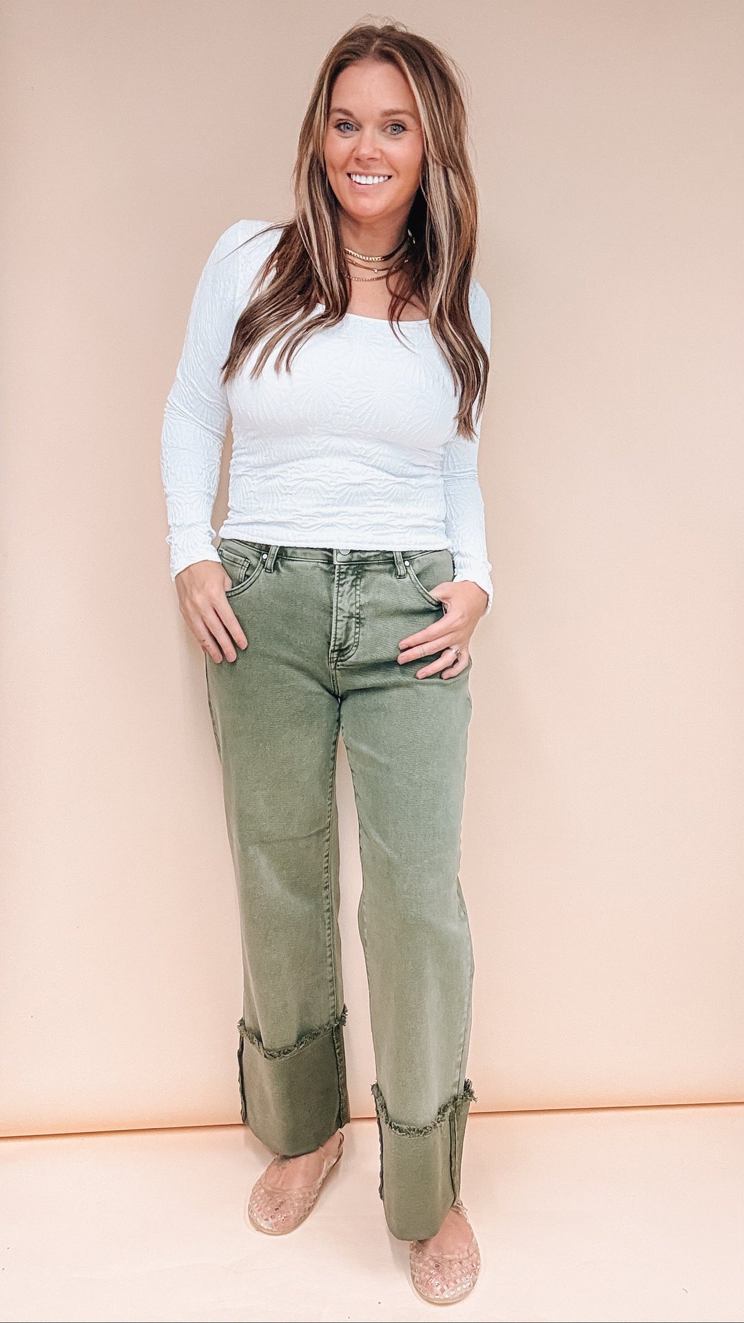 Woman wearing olive colored jeans and white long sleeve tee on plain background