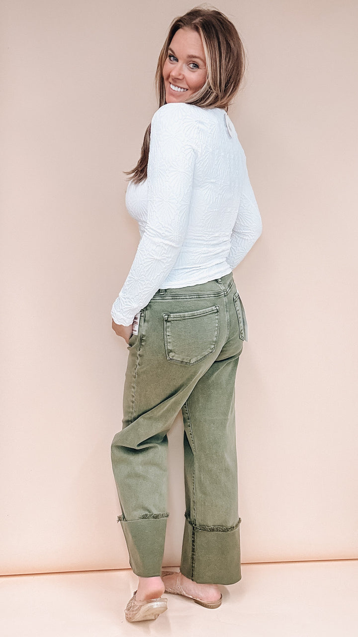 A woman wearing a white ribbed long-sleeve top and olive green wide-leg cropped pants, photographed from the back against a solid pale peach studio backdrop.