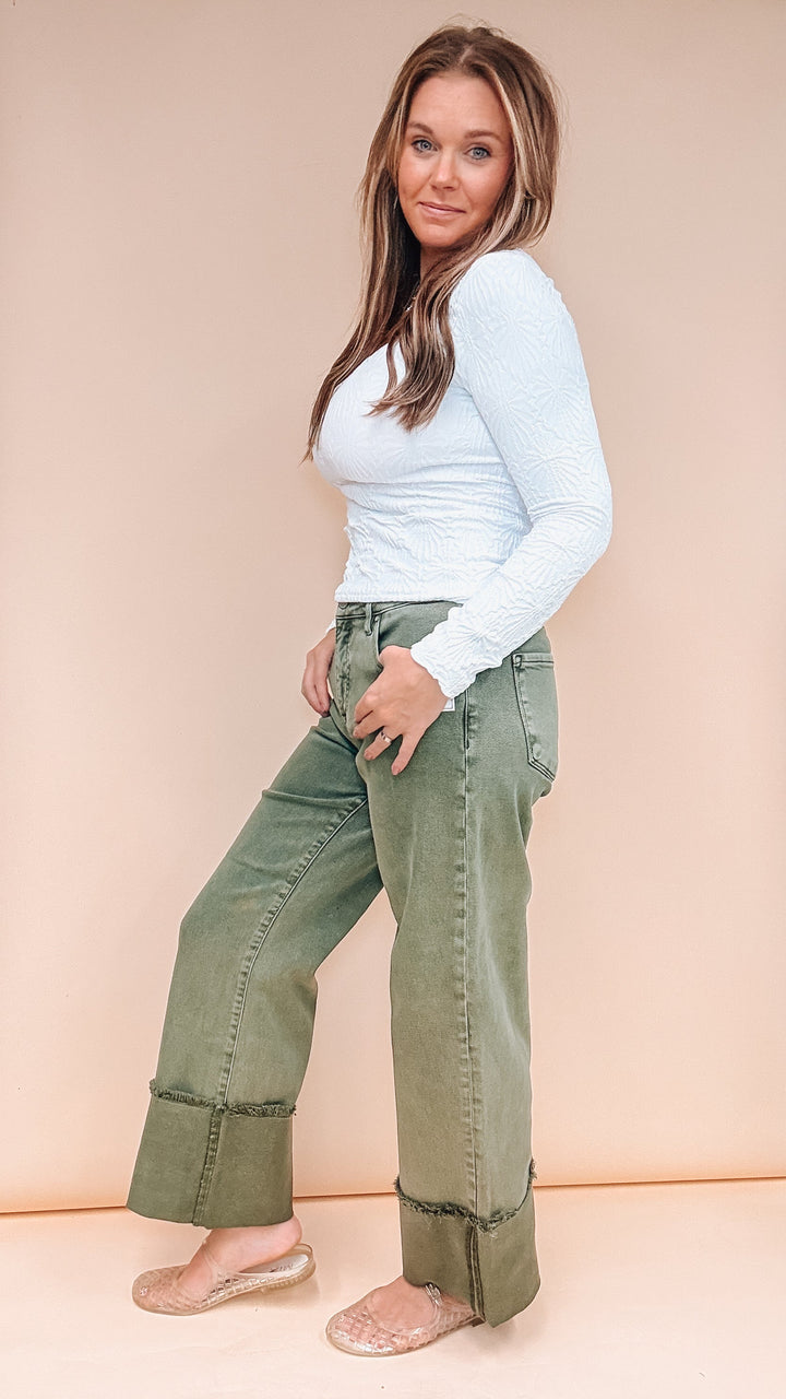 A woman modeling a pair of wide-leg, cuffed olive green jeans with a light blue long-sleeve top, standing against a solid peach-colored studio background.