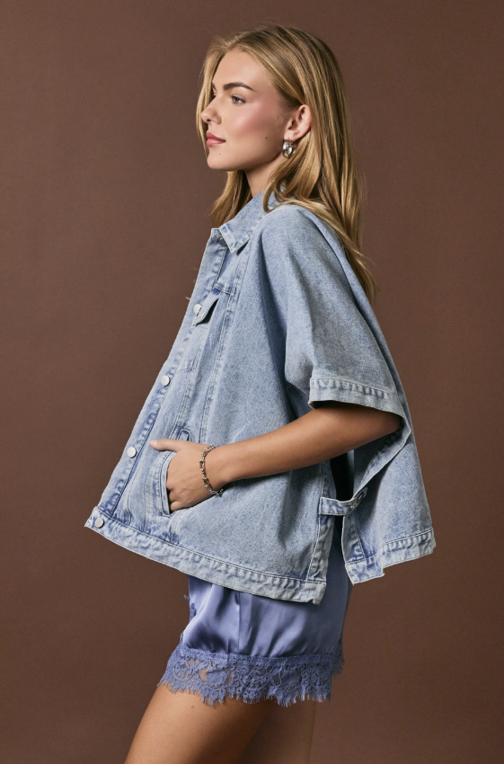 A woman wearing a light blue oversized denim jacket with wide short sleeves and front pockets, paired with matching light blue fringed shorts, posed against a solid brown studio background.