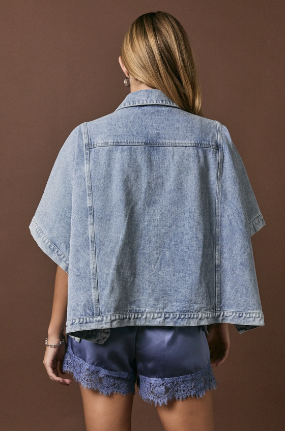 A woman shown from the back wearing a light-wash denim short-sleeve cropped jacket and dark blue lace-trim shorts against a solid brown studio background.
