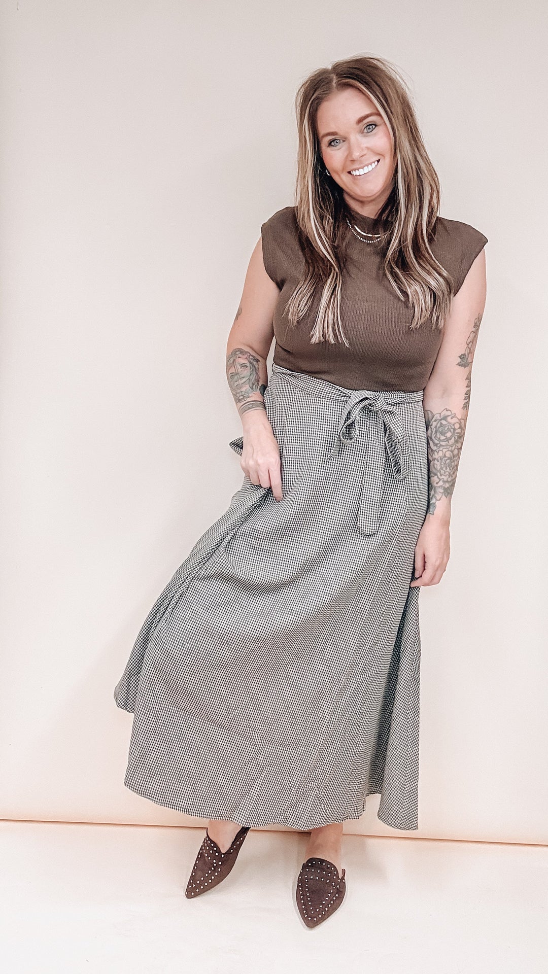 A woman modeling a brown sleeveless mock-neck top and a long gray wrap skirt with a tie waist, paired with brown pointed-toe flats, against a plain beige studio background.