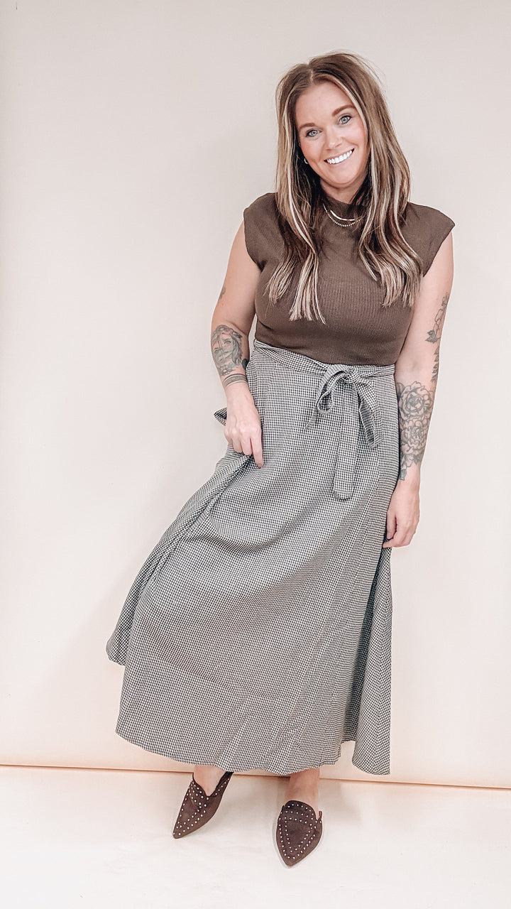 A woman modeling a brown sleeveless mock-neck top and a long gray wrap skirt with a tie waist, paired with brown pointed-toe flats, against a plain beige studio background.