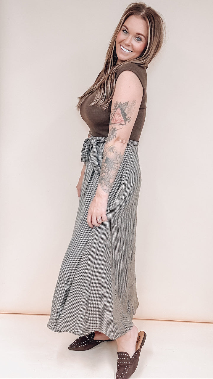 A woman modeling a sleeveless brown top and a long gray tie-waist skirt, paired with brown studded loafers, against a plain cream background.