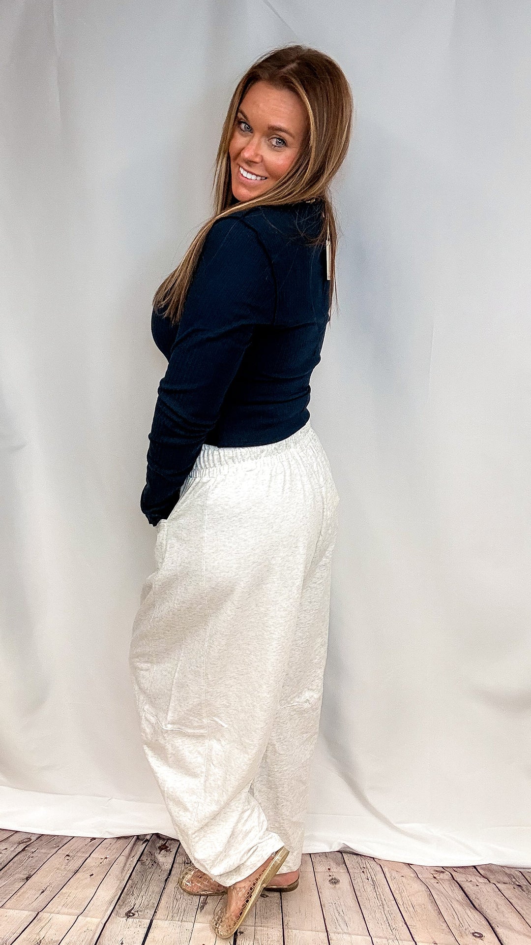 A woman posing in a dark navy long-sleeve top and textured cream wide-leg pants against a white draped studio backdrop.