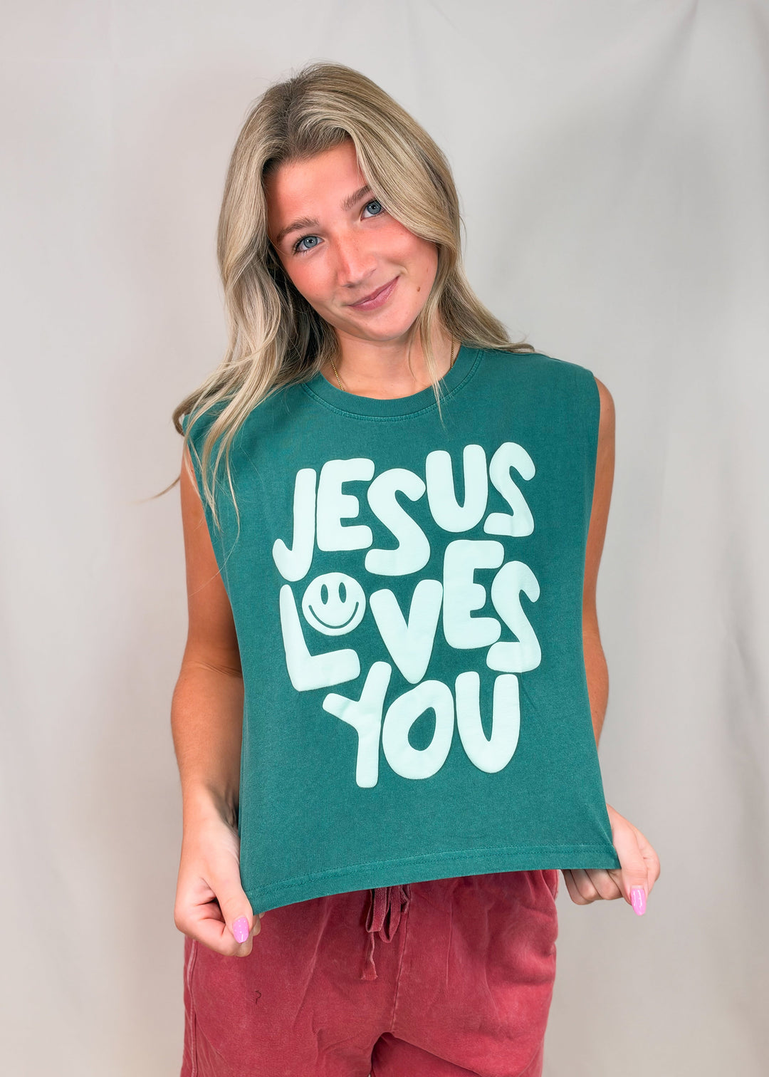 Woman wearing teal muscle tee with 'JESUS LOVES YOU' on front with red pants on plain background