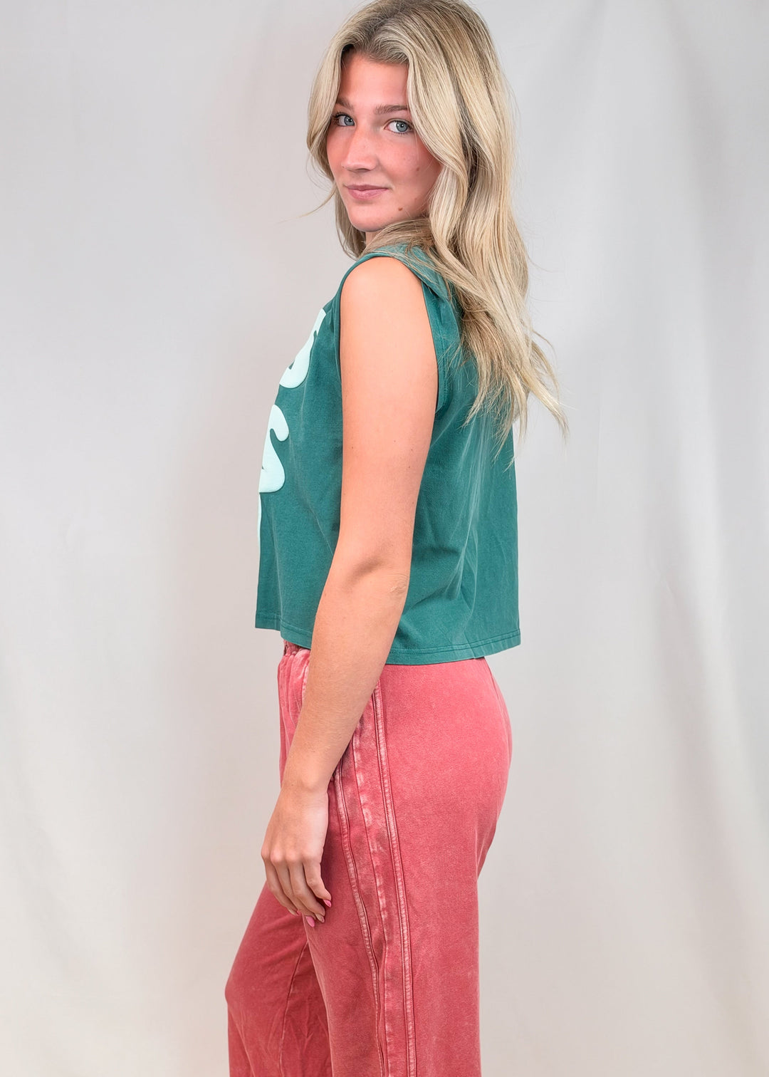 Woman shown from side view wearing teal muscle tee and red pants on plain background