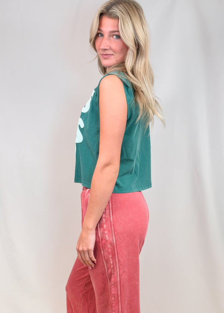 Woman shown from side view wearing teal muscle tee and red pants on plain background