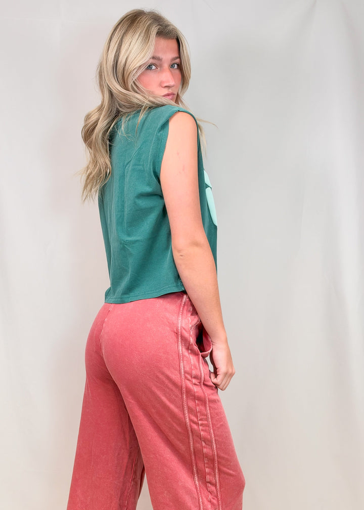 Woman shown from side view wearing teal muscle tank and red pants on plain background