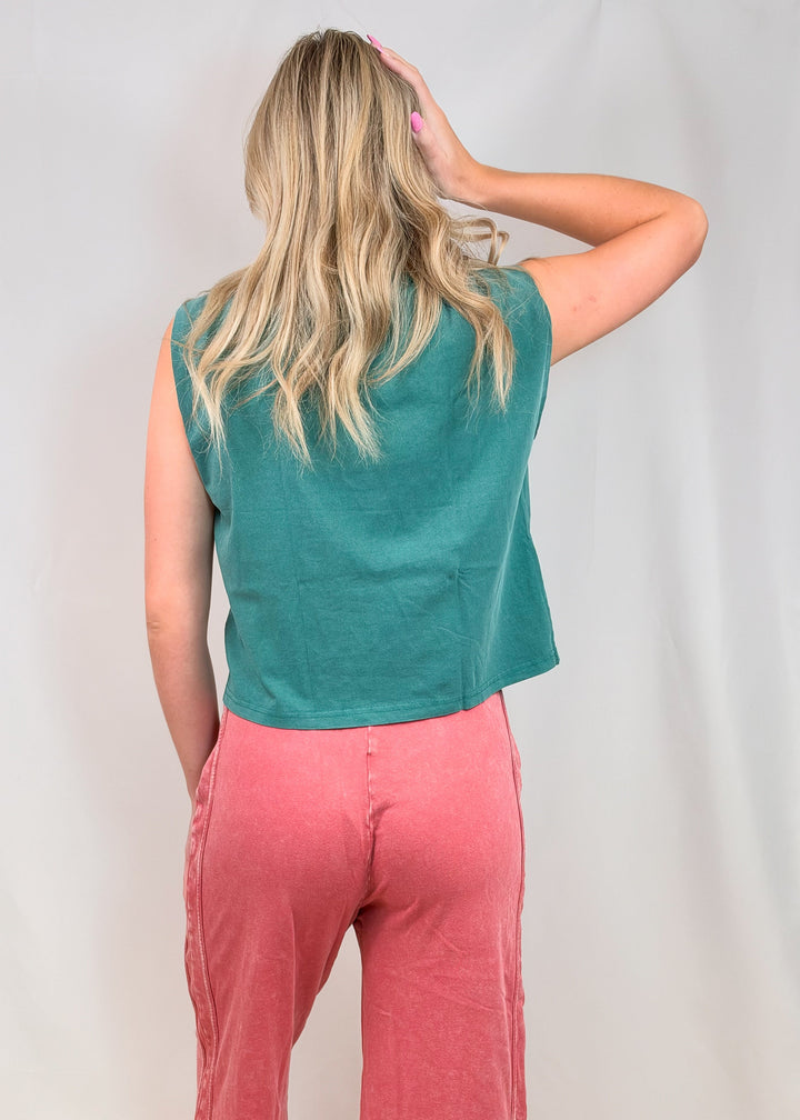 Woman shown from back view wearing teal muscle tank and red pants on plain background