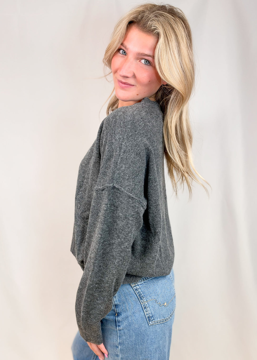 Woman shown from side view wearing charcoal grey sweater and jeans on plain background