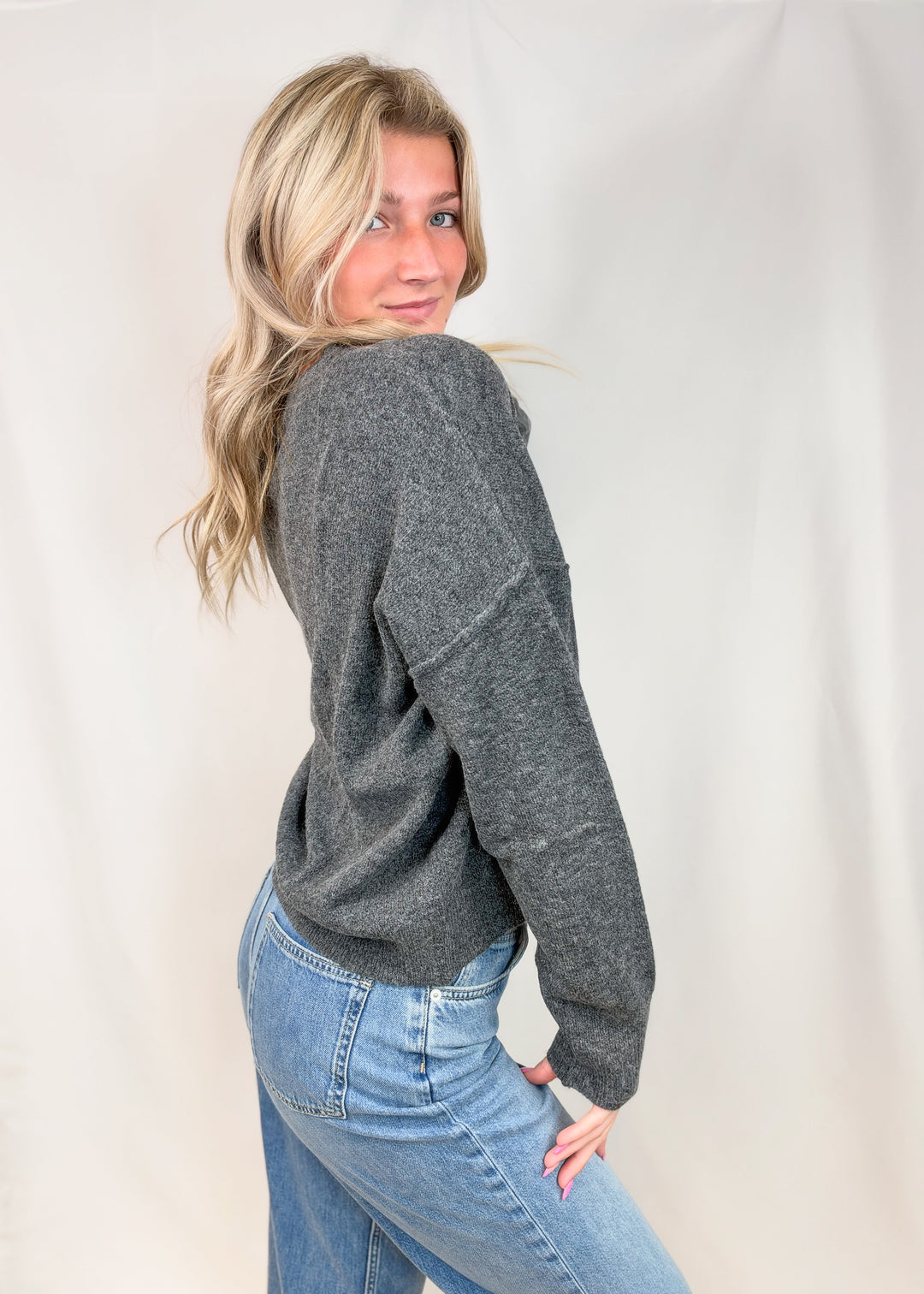 Woman shown from side view wearing charcoal grey sweater with jeans on plain background