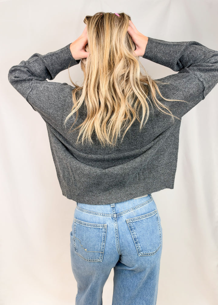 Woman shown from back view wearing charcoal grey sweater with jeans on plain background