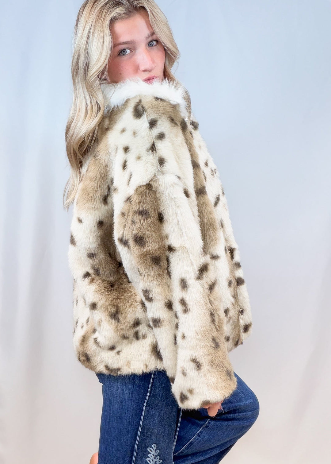 Woman shown from side view wearing faux fur animal print jacket with denim jeans on plain background