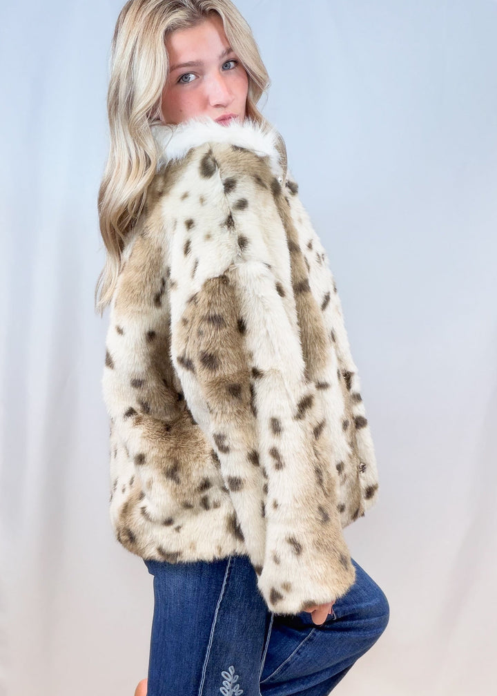 Woman shown from side view wearing faux fur animal print jacket with denim jeans on plain background