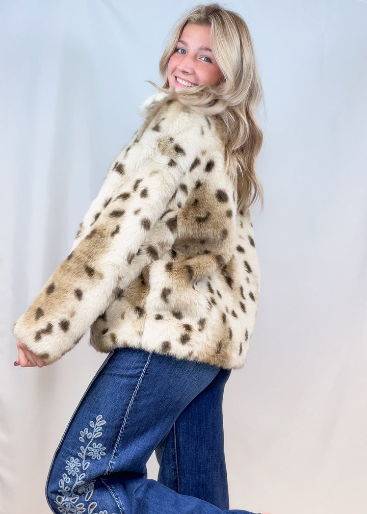 Woman shown from side view wearing faux fur animal print jacket with denim pants on plain background