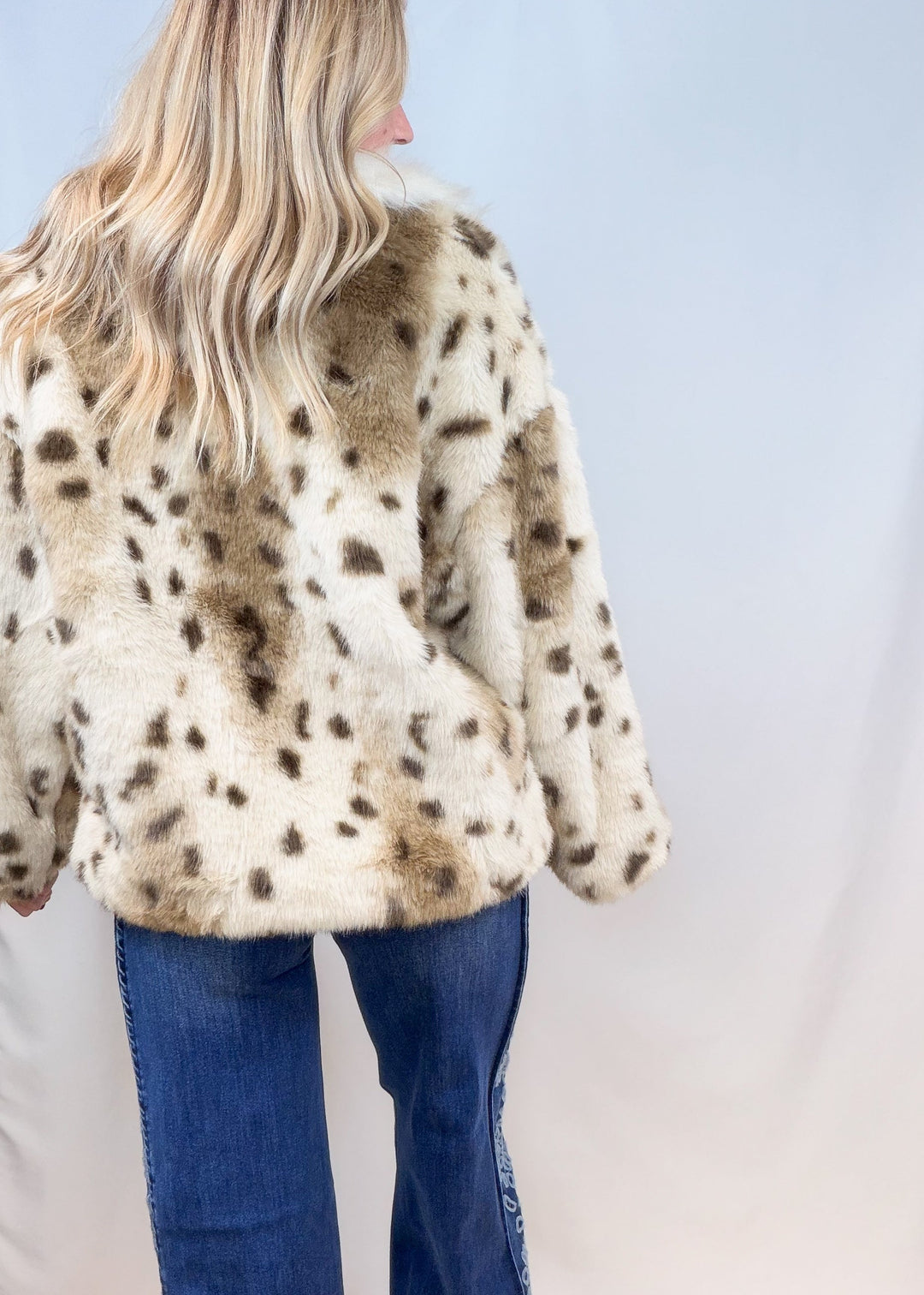 Woman shown from back view wearing faux fur animal print jacket with denim jeans on plain background