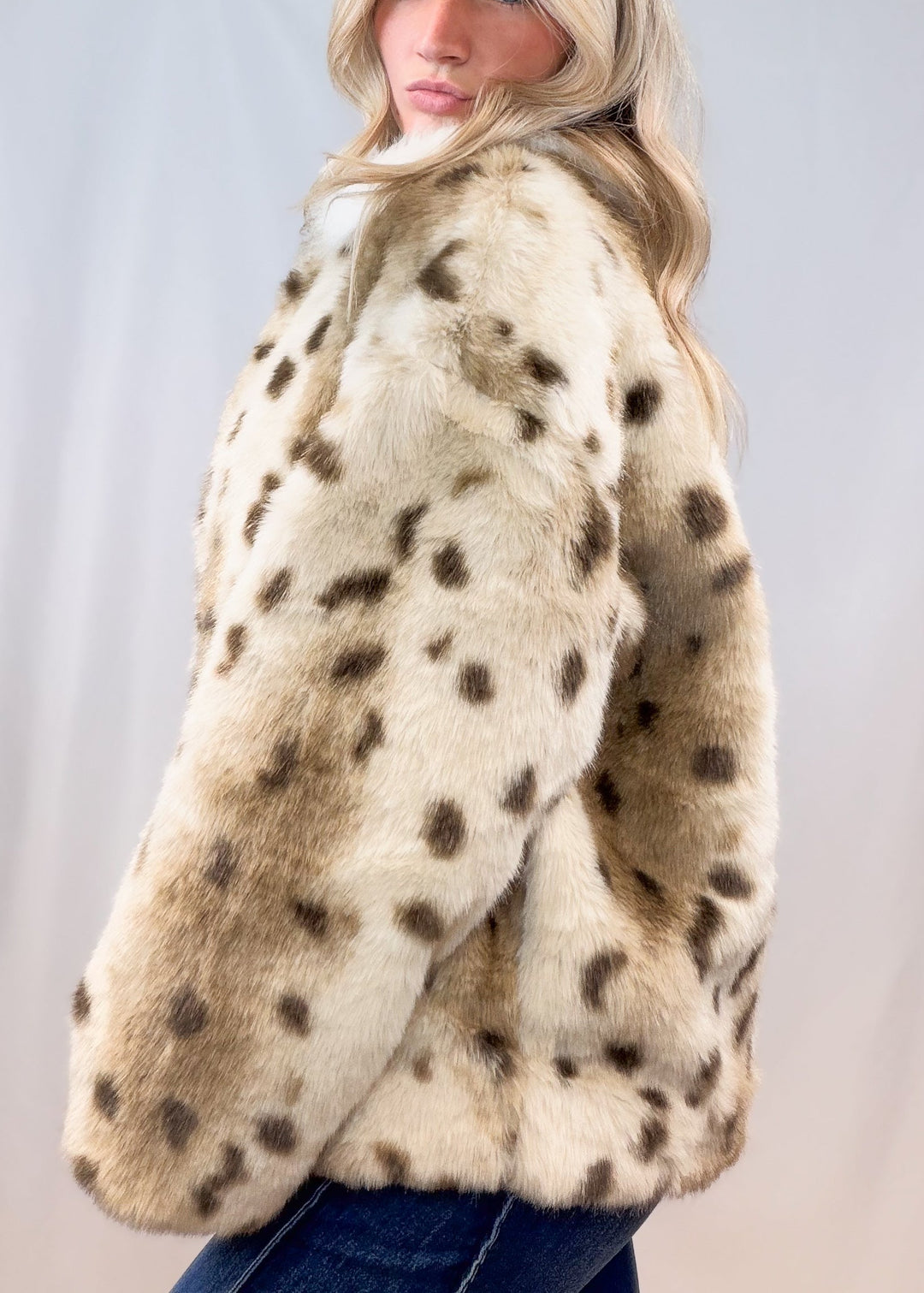 Woman shown from side view wearing faux fur animal print jacket with denim jeans on plain background
