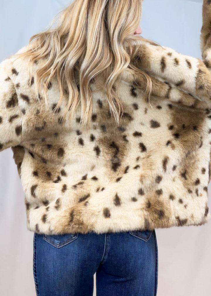 Woman shown from back view wearing faux fur animal print jacket with denim jeans on plain background