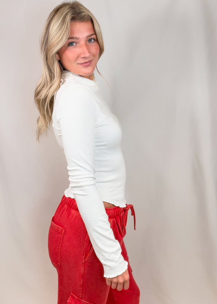 Woman shown from side view wearing white top with lettuce detail edges and red pants on plain background
