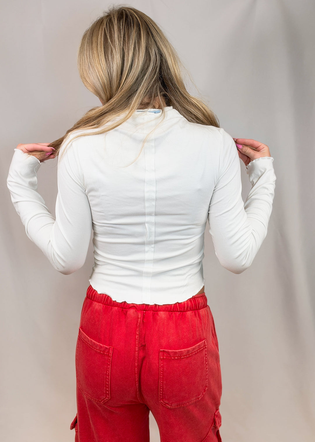 Woman shown from back view wearing white top with lettuce detail edges and red pants on plain background