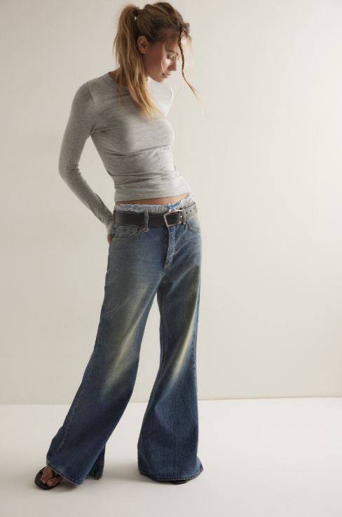 A woman modeling a light gray long-sleeve crop top and wide-leg blue jeans with a black belt, standing against a plain off-white studio background.