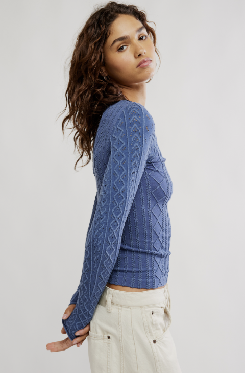 A woman wearing a blue cable-knit long-sleeve cropped top and cream pants, photographed against a plain light gray studio background.