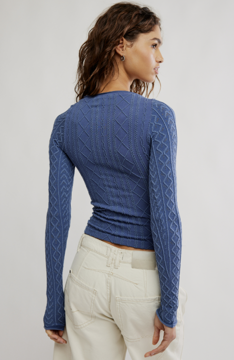 A woman shown from the back wearing a blue long-sleeve knit top with textured cable and diamond patterns, paired with off-white pants, against a light gray studio background.