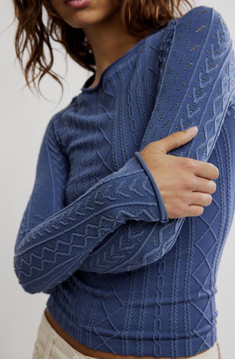 A close-up of a woman wearing a textured, long-sleeve knit top in a dusty medium blue. The top features intricate cable and eyelet knit patterns.